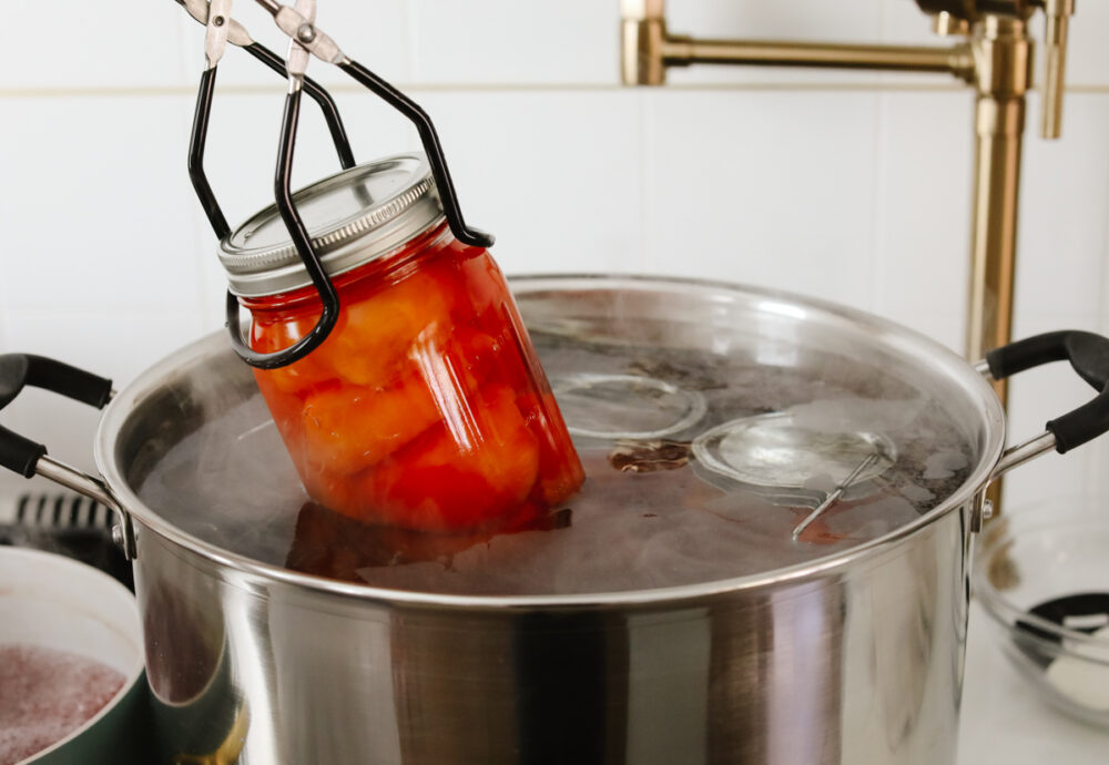 This in-depth guide to canning peaches gives you everything you need to know to have delicious peaches on hand year-round! It is easier than you would think and yields the sweetest, most delicious results each time. Adding a jar of canned peaches to boiling water for processing.