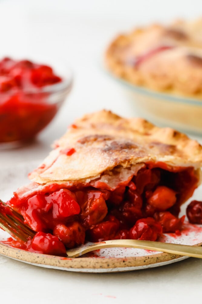 Homemade cherry pie is about as classic as they come and this recipe is no exception! Absolutely delicious, this mouthwatering pie has a tart filling and flaky crust that is so tasty that you won't be able to stop at just one slice! A slice of cherry pie on a plate with a fork.