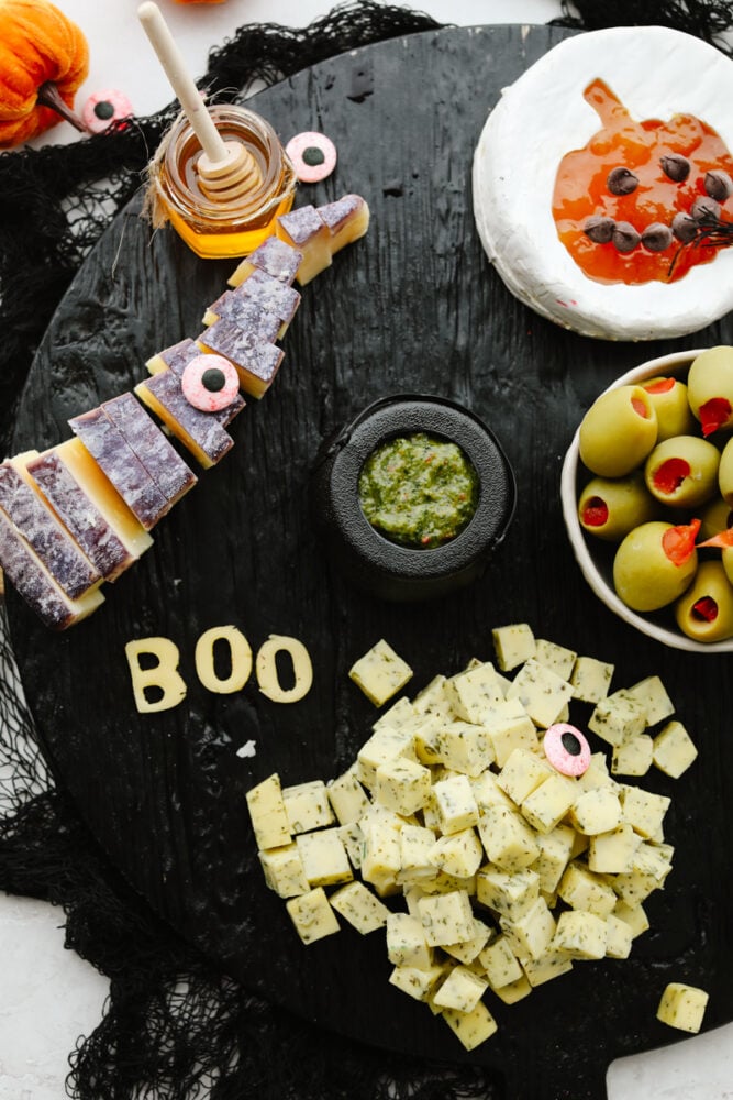 This Halloween charcuterie board is a ghoulish treat for any party! A spooky twist on your classic cheese board, it's filled with fruit, crackers, and meat and sure to be everyone's favorite! Spreads being added to a black board.