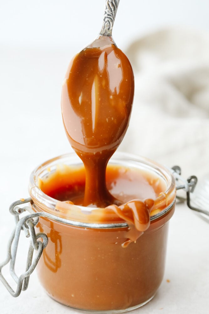 This homemade caramel sauce is straight from heaven! Its ooey-gooey, deliciously sweet flavor will have you over the moon excited, especially when you realize that you can add this to just about anything! Caramel being dipped into a jar with a spoon.