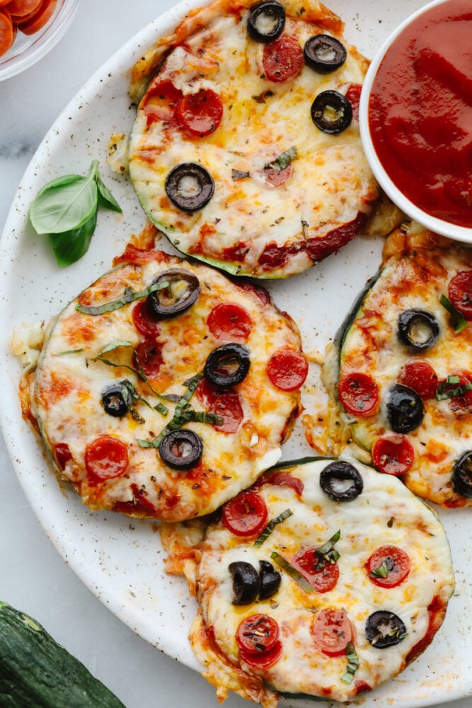 These melty, cheesy zucchini pizza bites have all the goodness of pizza without the carbs! In just 15 minutes, you have a tasty appetizer that you can feel great about eating! Pizza bites on a white plate with marinara dipping sauce.