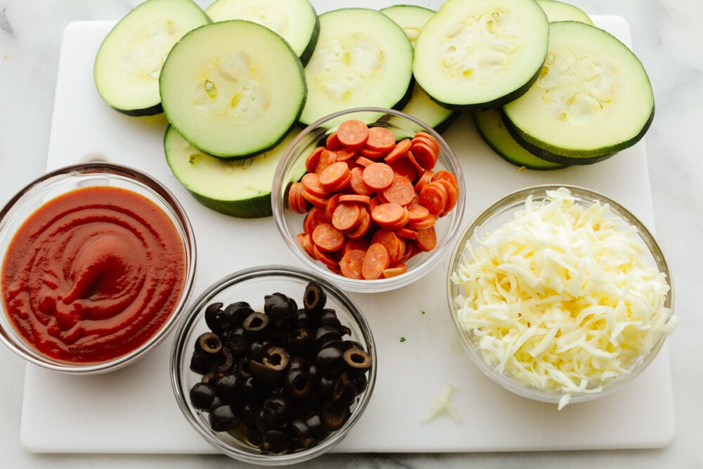 These melty, cheesy zucchini pizza bites have all the goodness of pizza without the carbs! In just 15 minutes, you have a tasty appetizer that you can feel great about eating! Zucchini pizza bite ingredients separated into small glass bowls.