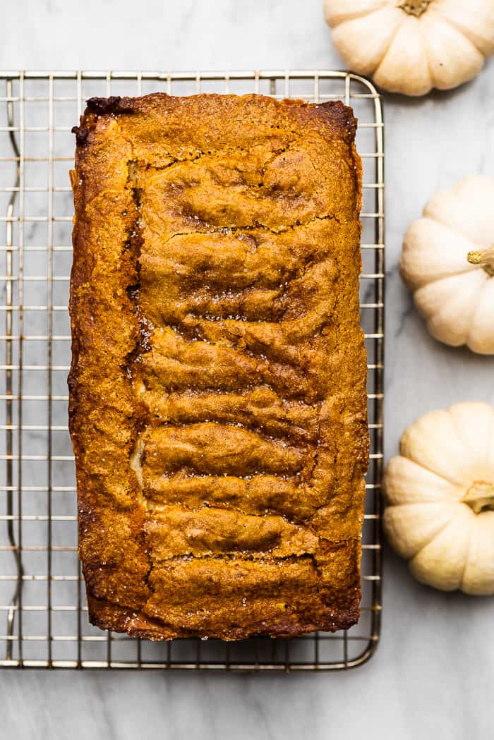 This cream cheese pumpkin bread recipe is truly the best ever. The moist and tender pumpkin bread is swirled with cream cheese frosting that's baked right into the loaf. It makes the perfect fall treat! cream cheese pumpkin loaf on a cooling rack surrounded by white pumpkins.
