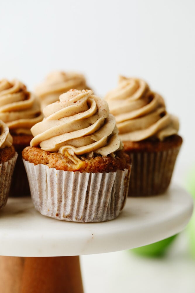 Apple pie cupcakes are a twist on a classic dessert. They are moist, delicious and with fresh apple pie filling in the middle, they give you JUST the right amount of texture! Close up of an apple pie cupcake on a cake stand.