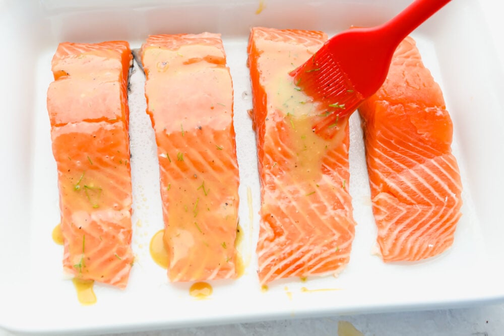 Honey mustard salmon is the perfect blend of tangy and delicious. It's tender and flaky and it completely falls apart on your fork! This tried and true recipe is one that your family will go crazy for! Salmon filets being basted before being cooked.