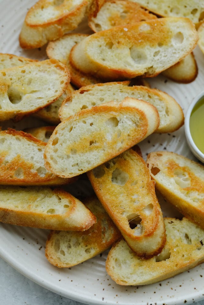 This easy, baked crostini is the best side to any Italian meal! They're golden, delicate pieces of heaven ready for your favorite toppings! Closeup of crostini pieces on a white plate.