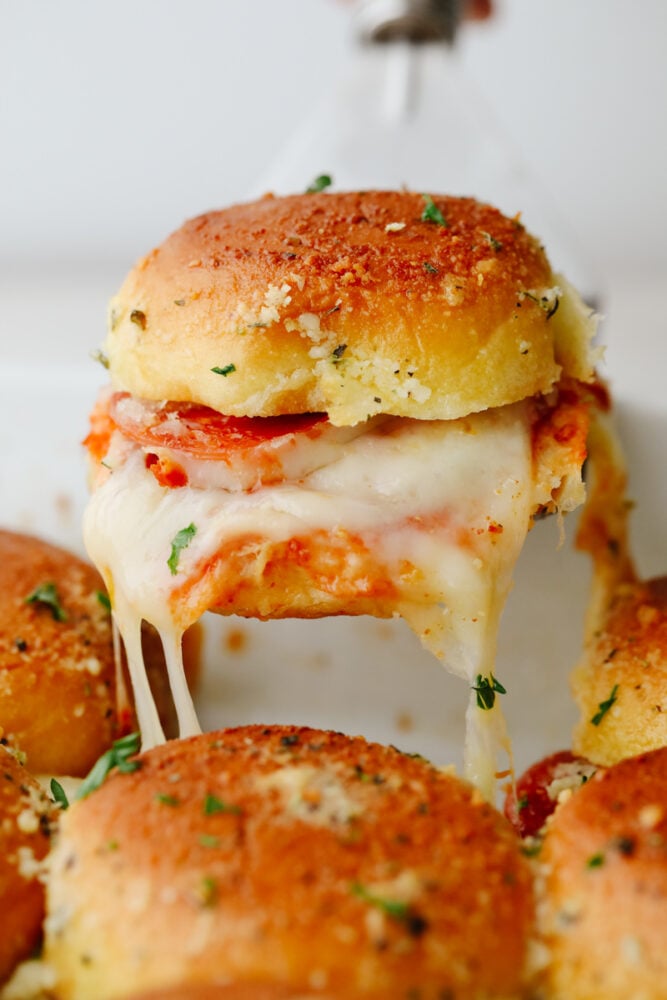 These pizza sliders are about to become your new favorite appetizer. Brushed with a buttery Italian seasoning glaze and filled with all of your favorite pizza toppings, each bite is truly irresistible! Closeup of pulling a pizza slider out of the pan.