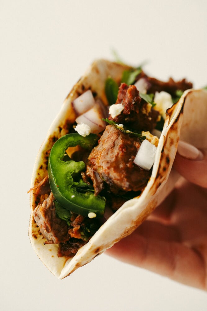Beef brisket tacos are full of delicious flavor and topped with perfectly tender and juicy meat. This is a meal that your entire family will go crazy over! A close up of a beef brisket taco folded in half being held in someone's hand.