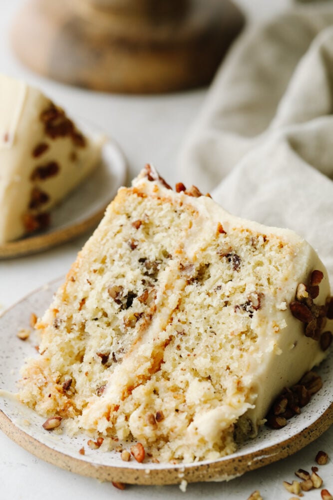 Soft, tender, and filled with nutty goodness, this butter pecan cake is perfect for fall! Browned butter cream cheese frosting and fresh chopped pecans make each bite a dream. A slice of butter pecan cake on a small stoneware dish.