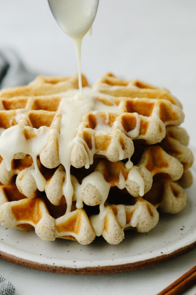 You just can't beat a delicious cinnamon waffle that is drizzled with a cream cheese glaze! It tastes just like a cinnamon roll in waffle form! A cinnamon roll waffle with cream cheese glaze being drizzled on it.