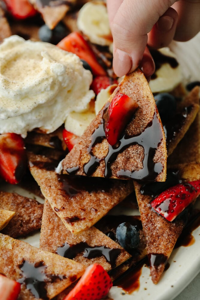 These dessert nachos are so delicious! Cinnamon sugar tortilla chips that are topped with fruit, chocolate and whipped cream... You won't find a dessert more loved than this one! Someone picking up a piece of dessert nachos.