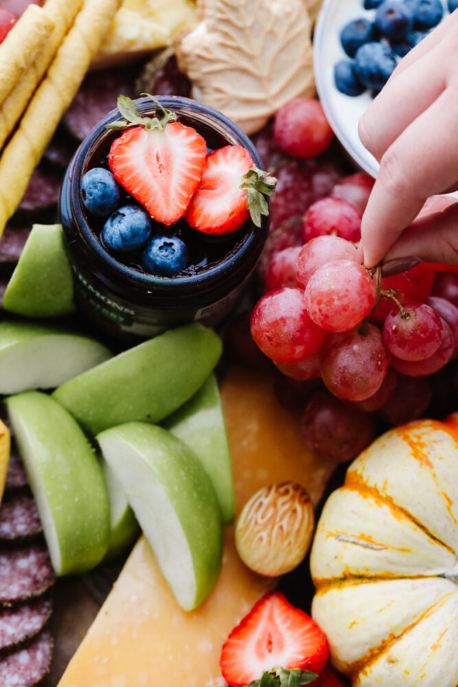 This fall charcuterie board is easy to make and is perfect for all of the parties this season! Packed with colorful and delicious food it is sure to be a hit with all of your guests! A hand picking up some fruit on a charcuterie board.
