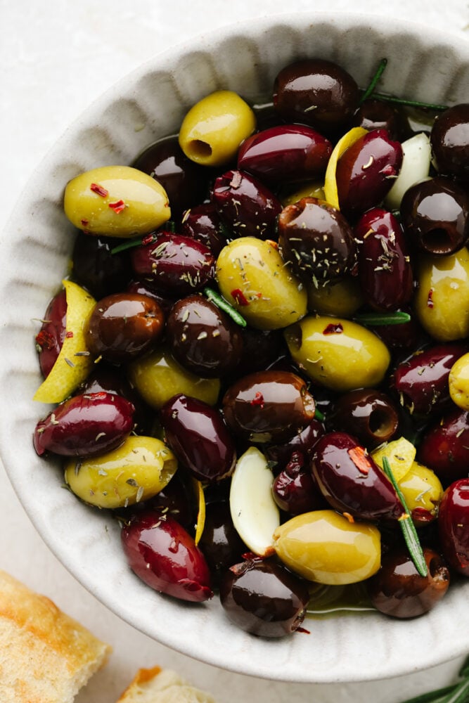Marinated olives is a fancy appetizer that comes together with little effort and inexpensive ingredients! Meaty, flavorful olives are layered with fresh herbs, spices, and olive oil for an experience your taste buds will absolutely love! Closeup of seasoned olives in a white bowl.