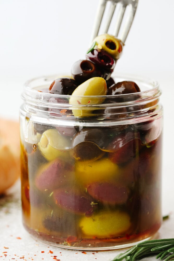 Marinated olives is a fancy appetizer that comes together with little effort and inexpensive ingredients! Meaty, flavorful olives are layered with fresh herbs, spices, and olive oil for an experience your taste buds will absolutely love! A jar of olives in marinade. Skewering 3 on a fork.
