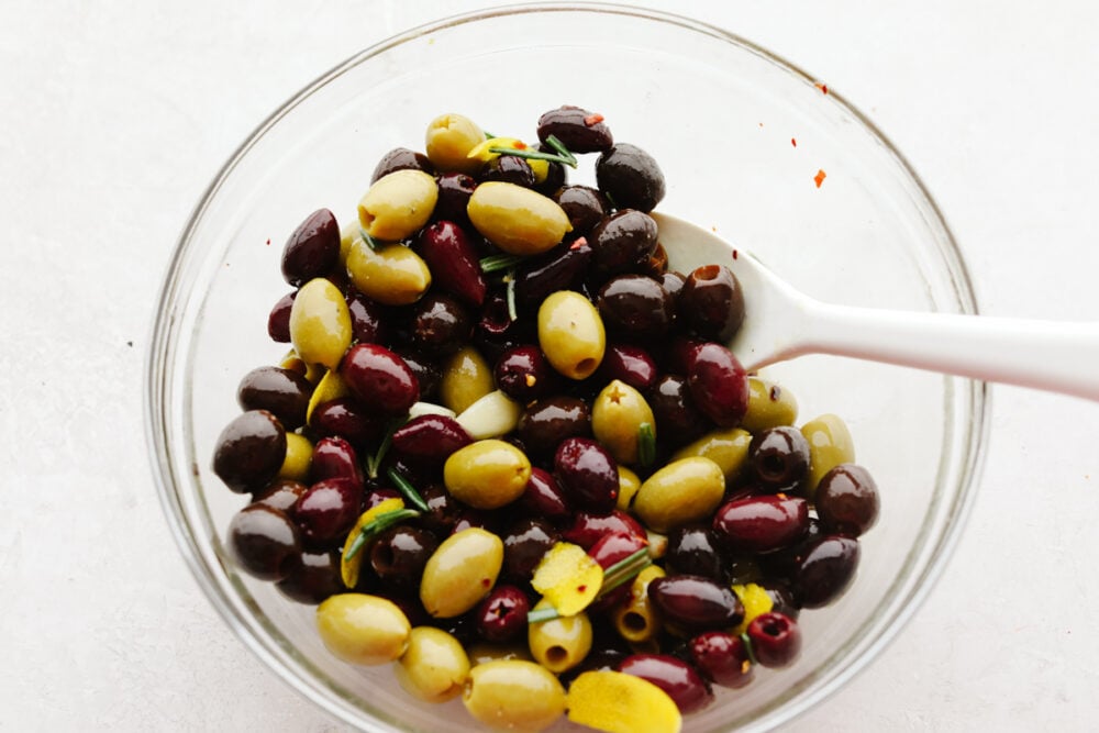 Marinated olives is a fancy appetizer that comes together with little effort and inexpensive ingredients! Meaty, flavorful olives are layered with fresh herbs, spices, and olive oil for an experience your taste buds will absolutely love! Mixing olives with herbs, lemon, and spices.