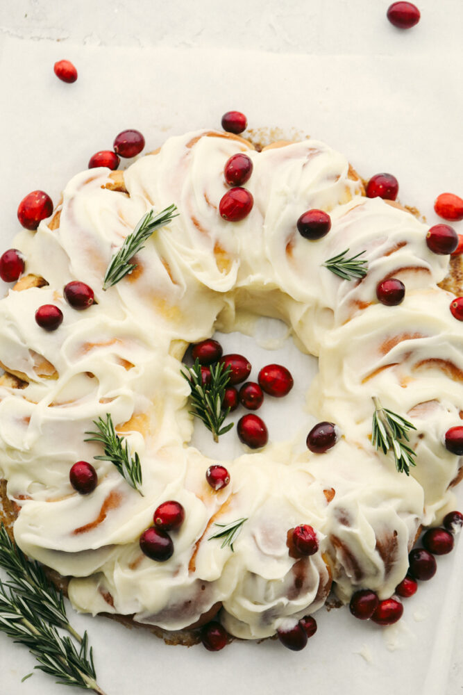 The holiday season doesn't get much more festive at the dinner table than with this cinnamon roll wreath! It's beautiful and delicious which makes it the perfect choice for your holiday breakfast, brunch, or dessert! A cinnamon roll wreath with cranberries and green garnish.