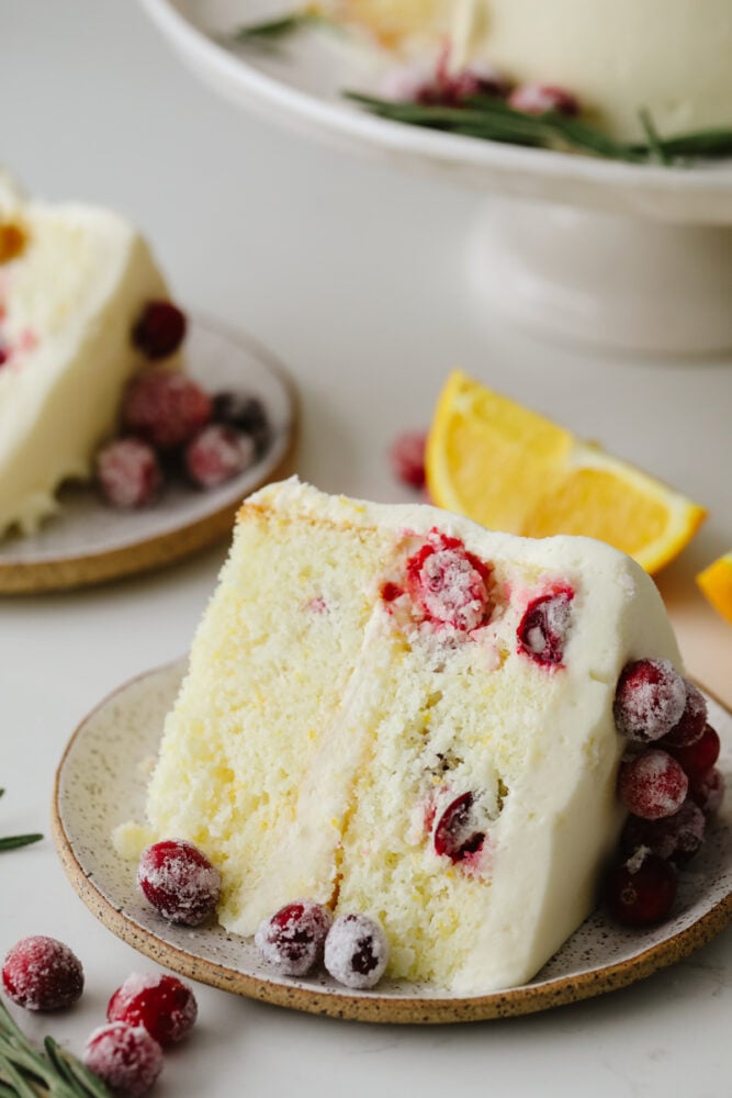 Cranberry Christmas cake is a show-stopping dessert you've got to try this holiday season! It's an amazing white cake filled with cranberries and topped with indulgent white chocolate buttercream! A slice of cranberry Christmas cake on a stoneware plate.