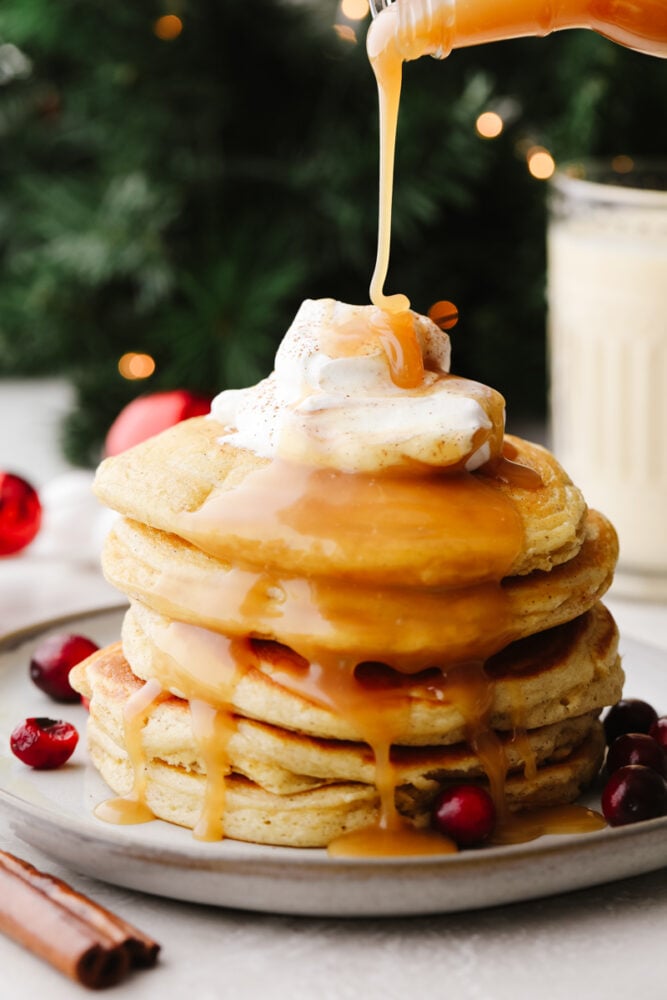 These eggnog pancakes are a MUST TRY this holiday season! They are soft and sweet and will melt right in your mouth. The hint of eggnog will have everyone coming back for more! Caramel sauce being drizzled onto a stack of eggnog pancakes.