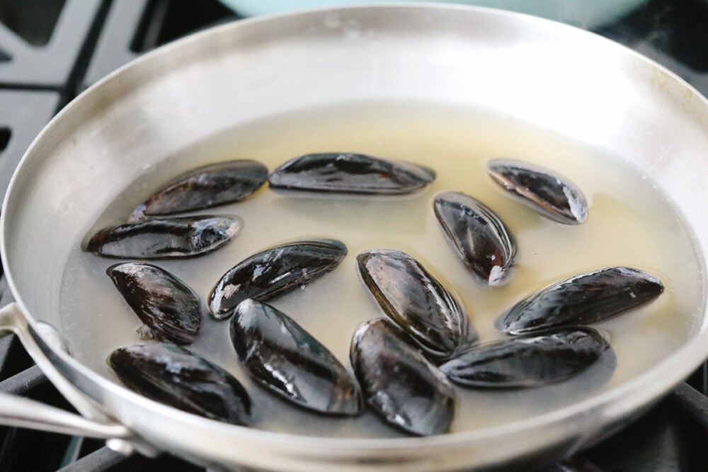 These steamed mussels are easy, elegant, and delicious! They are perfect for any dinner party and you won't be able to stop after just one! Uncooked mussels in a pan with white wine and chicken broth.