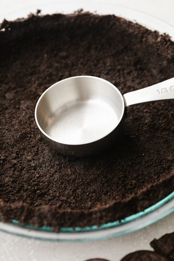 This Oreo pie crust is simple and delicious and it only takes 10 minutes to make! The chocolatey taste of Oreo's mixed with a creamy pie filling will make any pie taste SOO GOOD! A measuring cup pressing the Oreo crust down.