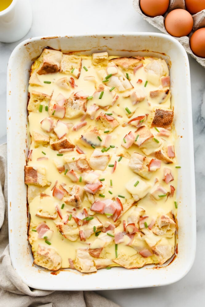 This overnight eggs benedict casserole is super easy to make and loaded with flavor! With layers of English muffins and Canadian bacon, all topped with a zesty hollandaise sauce, breakfast doesnโt get better than this! Eggs benedict casserole in a white serving dish.