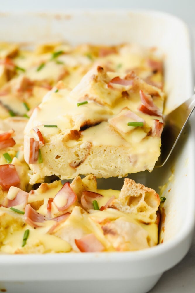 This overnight eggs benedict casserole is super easy to make and loaded with flavor! With layers of English muffins and Canadian bacon, all topped with a zesty hollandaise sauce, breakfast doesnโt get better than this! Taking a slice of casserole out of the pan.