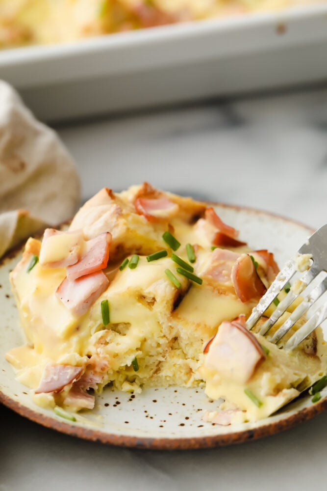 This overnight eggs benedict casserole is super easy to make and loaded with flavor! With layers of English muffins and Canadian bacon, all topped with a zesty hollandaise sauce, breakfast doesnโt get better than this! Taking a bite of egg benedict casserole topped with hollandaise sauce.