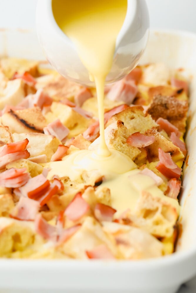 This overnight eggs benedict casserole is super easy to make and loaded with flavor! With layers of English muffins and Canadian bacon, all topped with a zesty hollandaise sauce, breakfast doesnโt get better than this! Pouring hollandaise sauce over casserole.