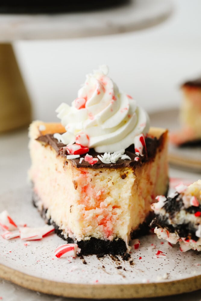 Peppermint cheesecake is creamy and delicious! It has the perfect oreo crust and is topped with crushed peppermint and whipped cream. This will definitely get a "WOW!" from all of your guests this holiday season! A slice of peppermint cheesecake on a plate with a bite taken out of it.