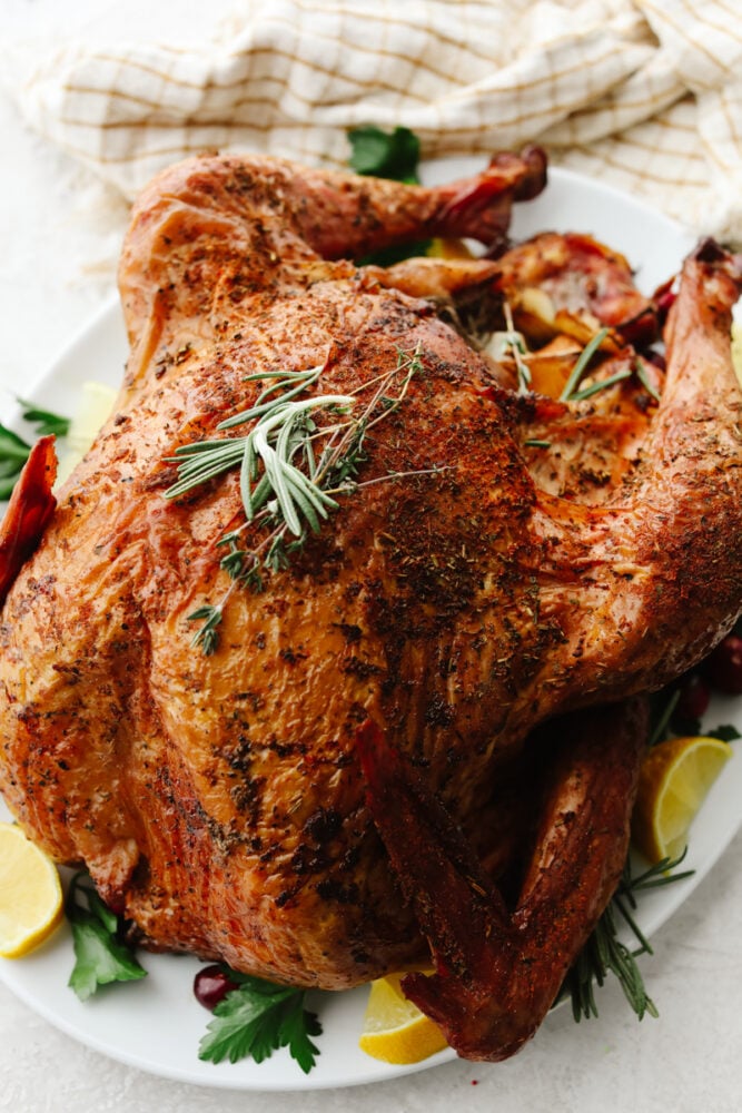 Just in time for Thanksgiving, this perfect smoked turkey is juicy and tender with a subtle smoky flavor. A simple brine and fresh herbs make each bite burst with flavor! A full smoked turkey on a white plate with garnish.