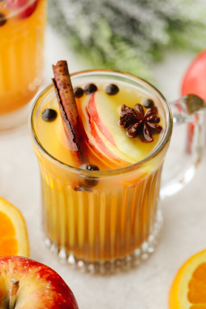 This traditional English drink is the perfect combination of fruit and spice! Wassail mixes up in a snap and is the perfect holiday crowd-pleaser! Wassail in a glass cup with a cinnamon stick and apple slices.