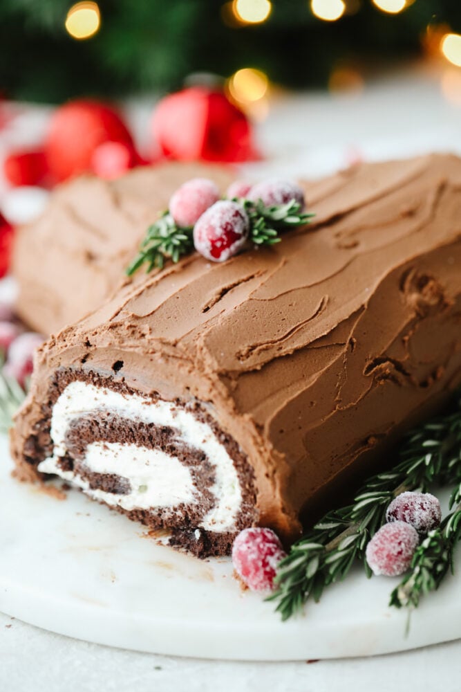 Show off your holiday spirit by baking this amazing yule log cake! A chocolate cake rolled with a cream cheese filling and topped with a chocolate ganache... It doesn't get much better than this! A yule log cake on a platter with garnish.