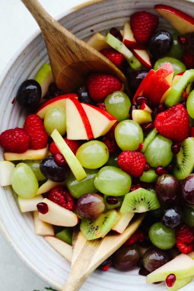 This easy Christmas fruit salad recipe is loaded with red and green fruit making it the perfect light and healthy addition to your holiday table! With a super simple dressing, it comes together quickly and is sure to be a hit! Christmas fruit salad in a bowl with a wooden spoon.