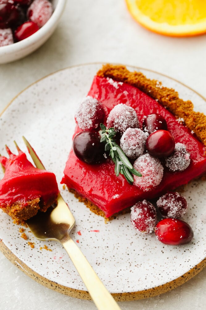 With a smooth tangy filling and Biscoff crust, this cranberry curd tart is a must-make for the holidays. It's as delicious as it is beautiful! A slice of cranberry curd tart on a plate with a fork bite.