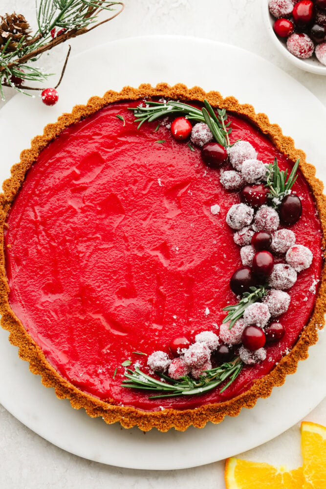 With a smooth tangy filling and Biscoff crust, this cranberry curd tart is a must-make for the holidays. It's as delicious as it is beautiful! A full cranberry curd tart, topped with sugared cranberries and rosemary garnish.