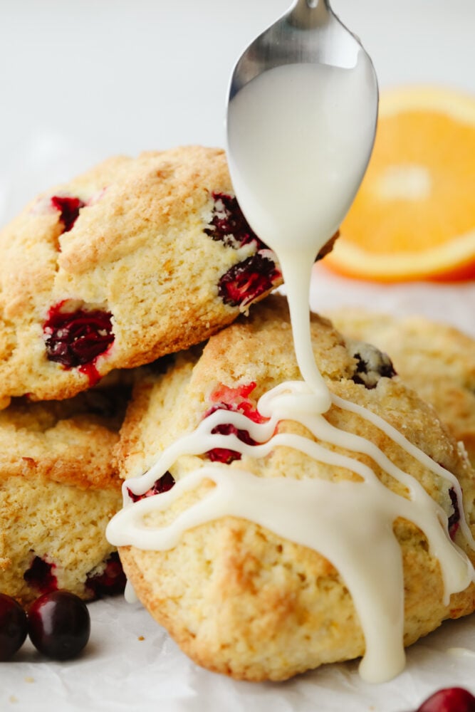 These cranberry orange scones are perfectly soft and fluffy on the inside and crisp and crunchy on the outside. They are bursting with cranberry and orange flavor and make the perfect breakfast! Orange glaze being drizzled on a scone.