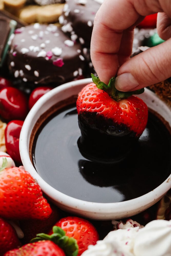 If you are looking for a Christmas dessert to impress your guests then this charcuterie board is it! Stunning and delicious, this board is full of candy, chocolate, cookies, and fruit for the holiday season!ย A strawberry being dipped into chocolate sauce.