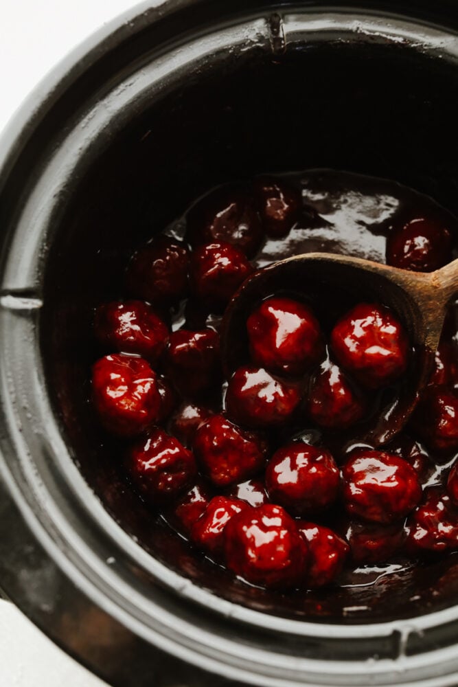 These grape jelly meatballs are so simple and easy and my whole family loves them! They pack so much flavor and are the perfect meal or appetizer for any occasion! A crockpot filled with meatballs and a wooden spoon.