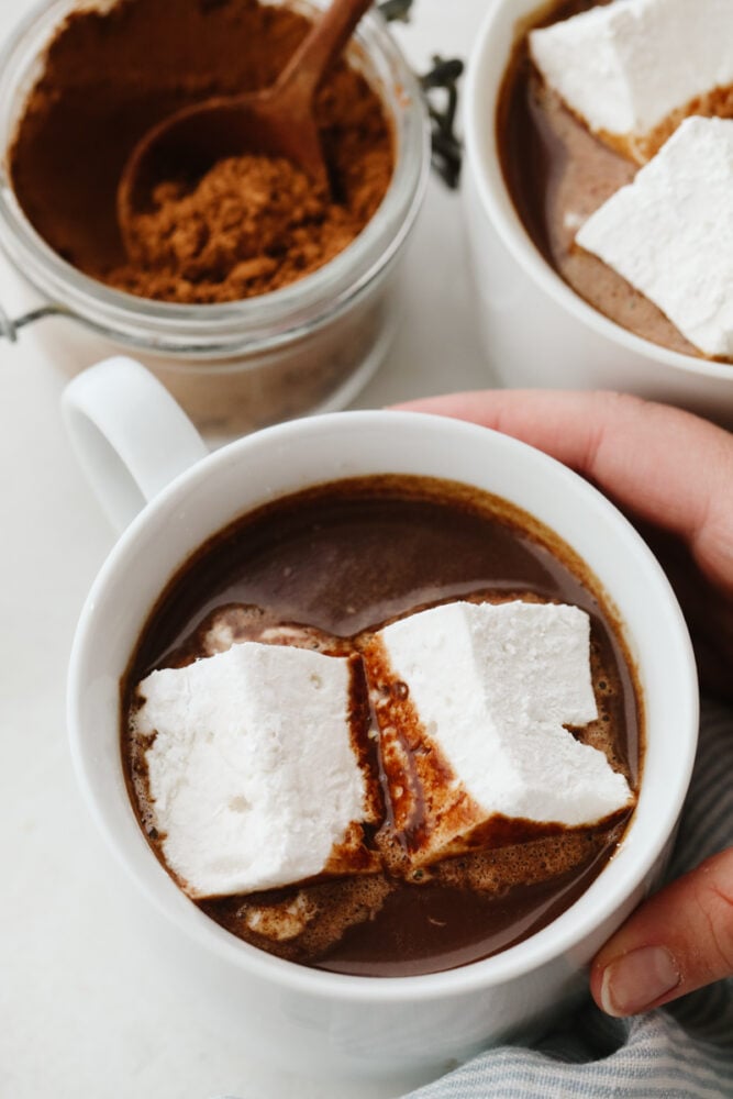 This easy homemade hot chocolate is the perfect treat to end your year with! It's so rich and creamy, you won't want it any other way! Holding a white mug filled with hot chocolate and marshmallows.