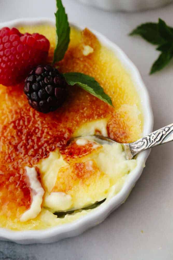 This creme brรปlรฉeย is a simple but elegant dessert that will impress all of your guests! You just can't beat a creamy custard topped with caramelized sugar and delicious fresh fruit! Creme brรปlรฉe in a ramekin with a spoon scooping some out.