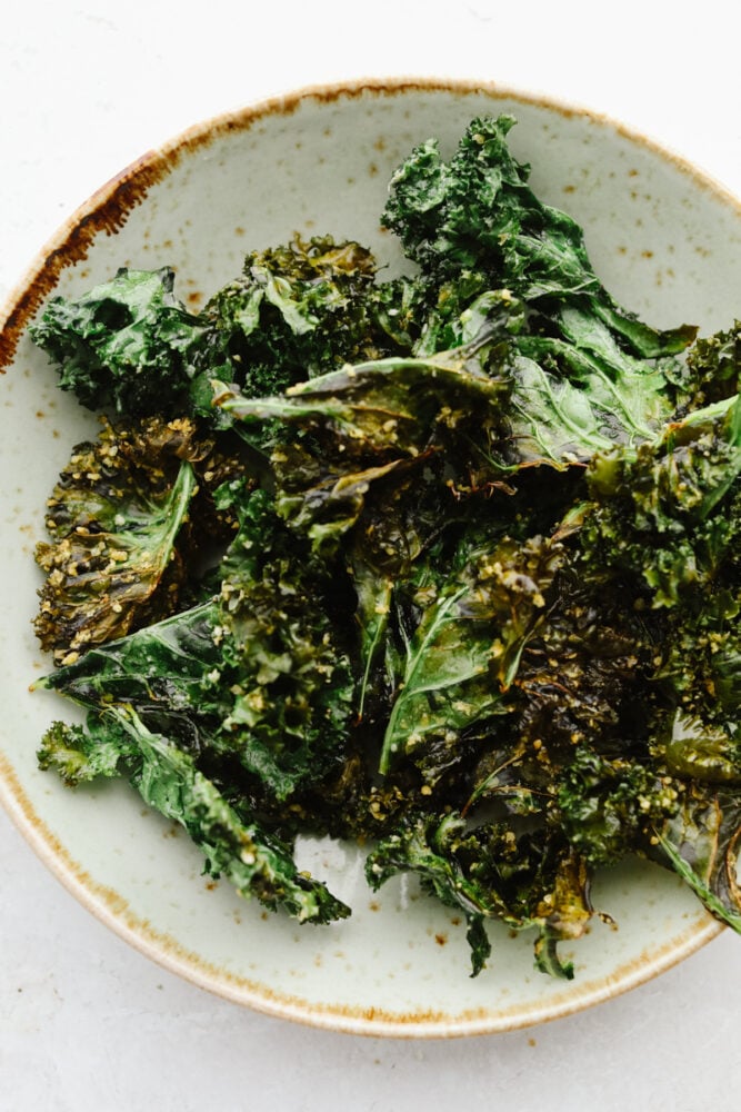 Crispy and seasoned perfectly with parmesan cheese, these kale chips that you cook in the air fryer are the BEST snack. They are quick and easy to make and I bet that you can't stop at just one! Cooked kale chips on a plate.