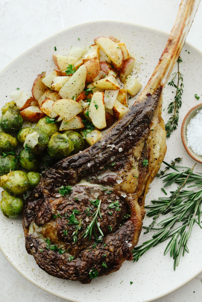 This tomahawk steak is juicy, tender, and filled with insanely delicious flavor! Make it your main dish for any occasion and your guests will keep coming back for more! A plate with steak, potatoes and brussel sprouts.
