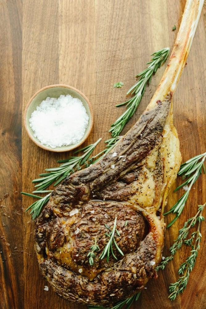 This tomahawk steak is juicy, tender, and filled with insanely delicious flavor! Make it your main dish for any occasion and your guests will keep coming back for more! A tomahawk steak with garnish on a cutting board.