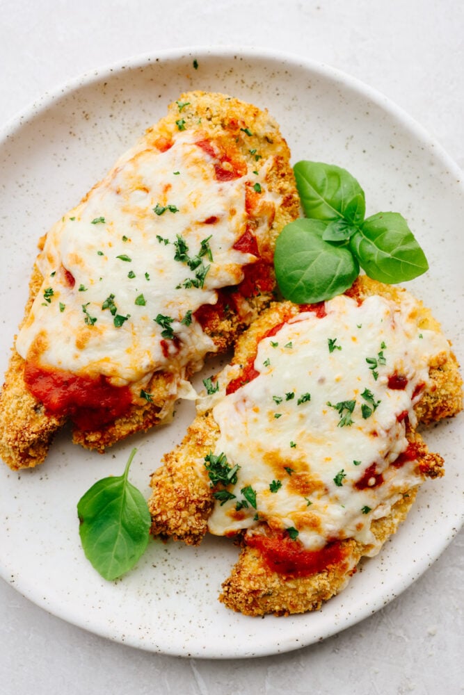 Air fryer chicken parmesan is cheesy, crispy, and cooks up in less than 10 minutes! It’s an easy-to-make dinner that your family will go crazy over! 2 pieces of air fryer chicken parmesan with herb garnish on a white plate.