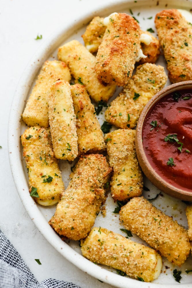 Crispy, cheesy, and delicious, these easy air fryer mozzarella sticks are going to be your new favorite snack! They're super simple to prepare and cook up in less than 10 minutes! Air fryer mozzarella sticks on a white plate served with marinara sauce.