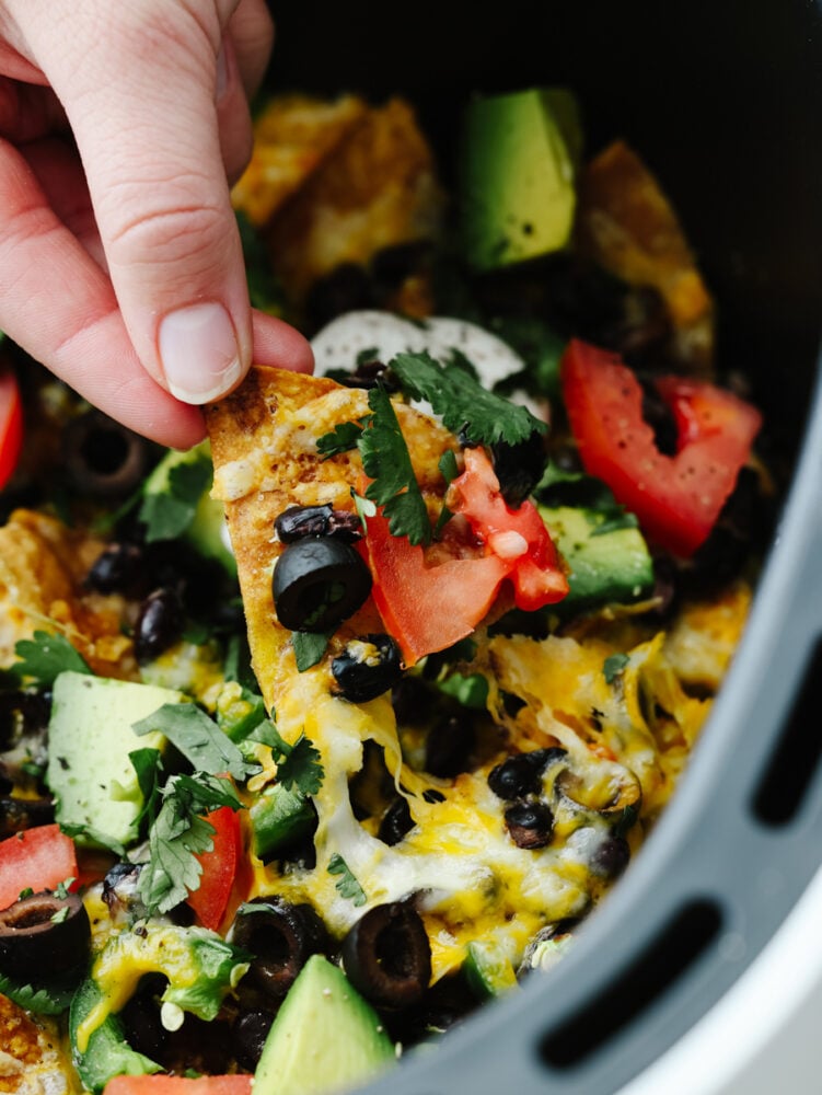 Air fryer nachos are loaded with cheese and vegetables and cooked to absolute perfection. This quick and easy appetizer is perfect for game day or any family get-together! A hand scooping a nacho out of an air fryer.