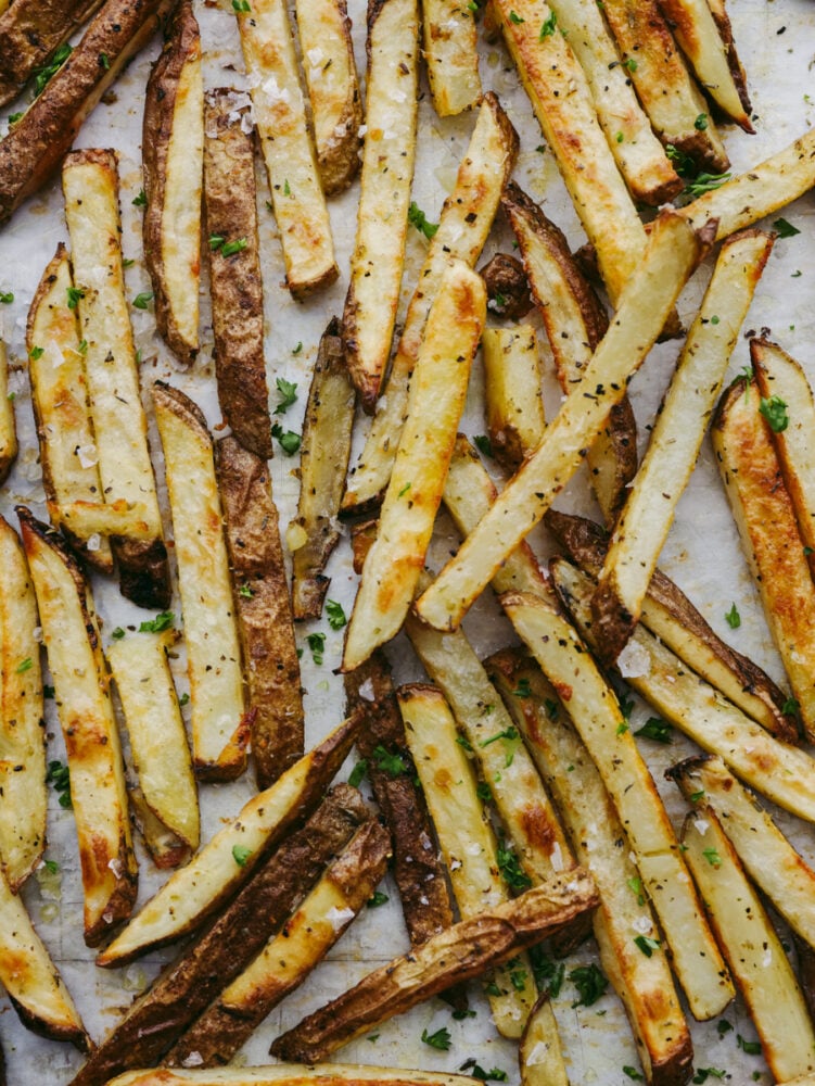 These golden, baked fries are perfectly crispy without the mess of deep-frying! Tossed with oil and tasty Italian herbs, these are sure to become a new family favorite! Baked fries topped with herbs laid out on parchment paper.