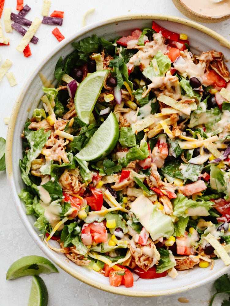 This BBQ chicken salad is healthy and packed to the brim with flavor! Filled with veggies, shredded chicken, and crispy tortilla chips, this salad really takes off when you add in the dressing. It's to die for! BBQ chicken salad with dressing and topped with fresh limes.