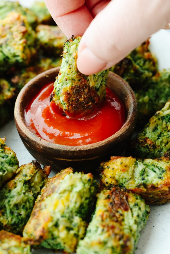 A delicious and cheesy twist on a classic favorite, these broccoli tots will fly off of your table. They are the perfect side or appetizer for any meal! A broccoli tot being dipped into ketchup.