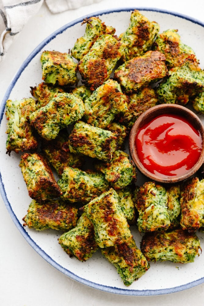 A delicious and cheesy twist on a classic favorite, these broccoli tots will fly off of your table. They are the perfect side or appetizer for any meal! A plate of broccoli too with some ketchup.